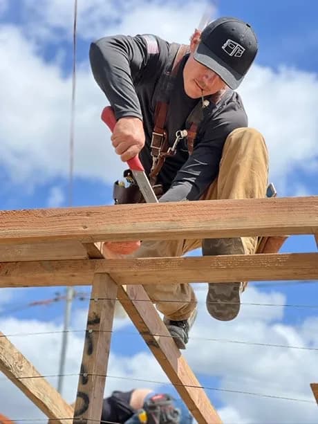 Luke Richard cutting a rafter by hand on top of a timber frame against a blue sky